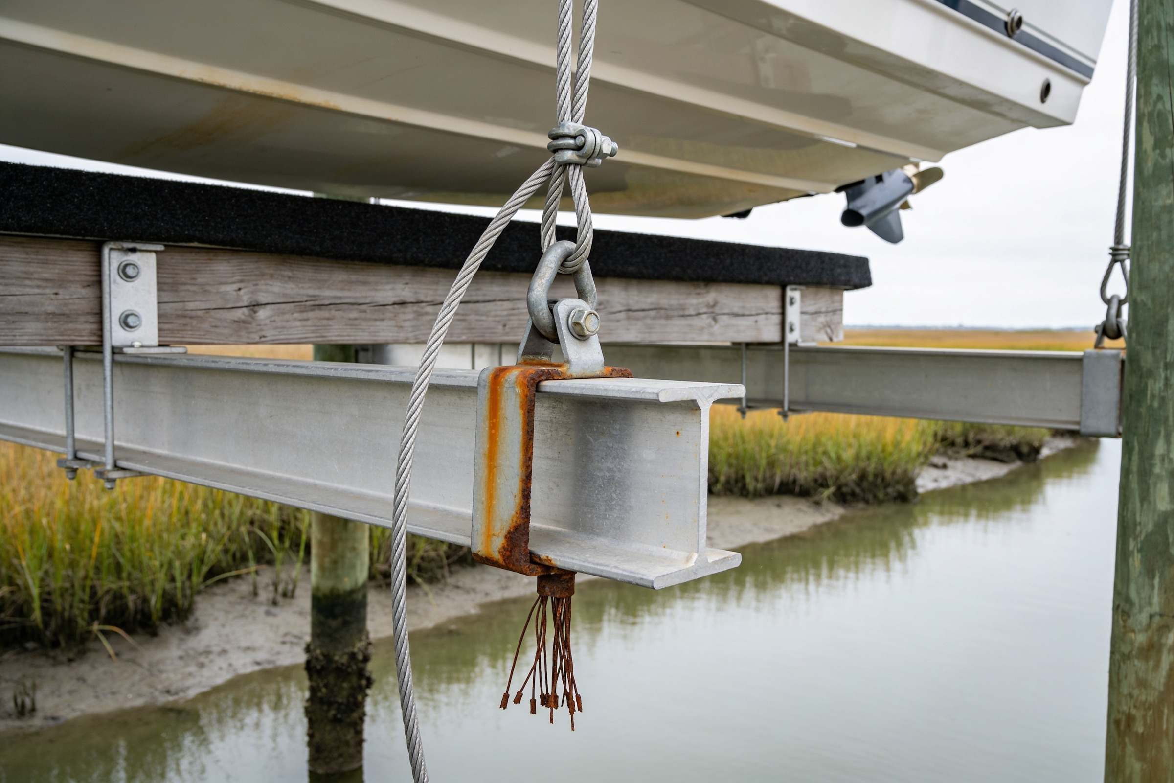 A boat lift cable fixed to the lift's aluminum beam by a rusted steel bracket, with the broken wire strands of the cable end hanging down from the bracket, rust-brown.