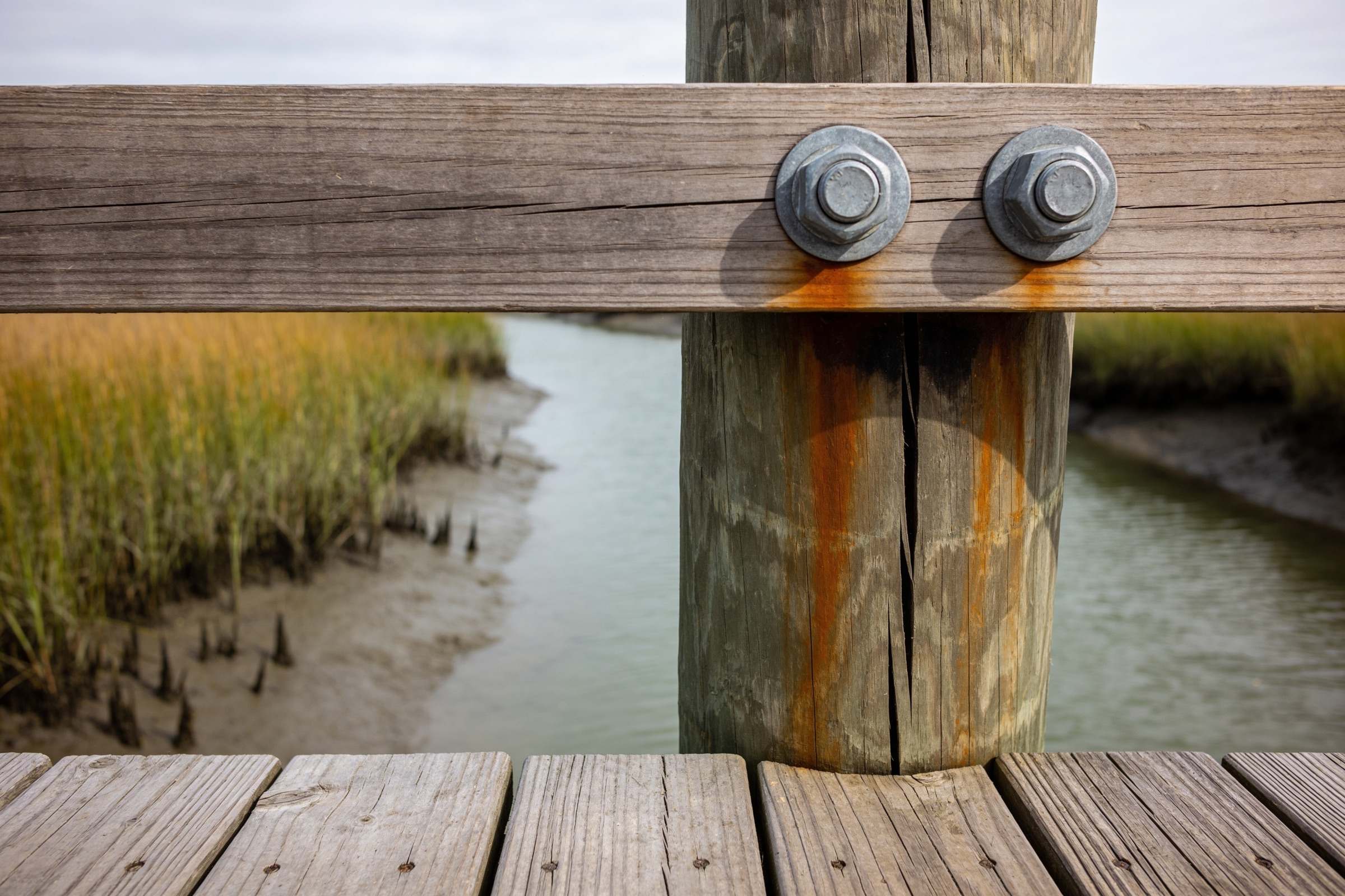 Two galvanized bolts through a horizontal beam into a dock piling, with orange rust running down the piling and a deep crack splitting the wood below the beam.
